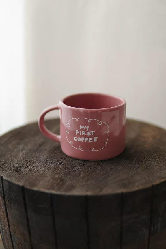 Mug My First Coffee en céramique rose de 2,5 dcl avec inscription blanche, posé sur un tonneau en bois rustique. Idéal pour le café du matin.