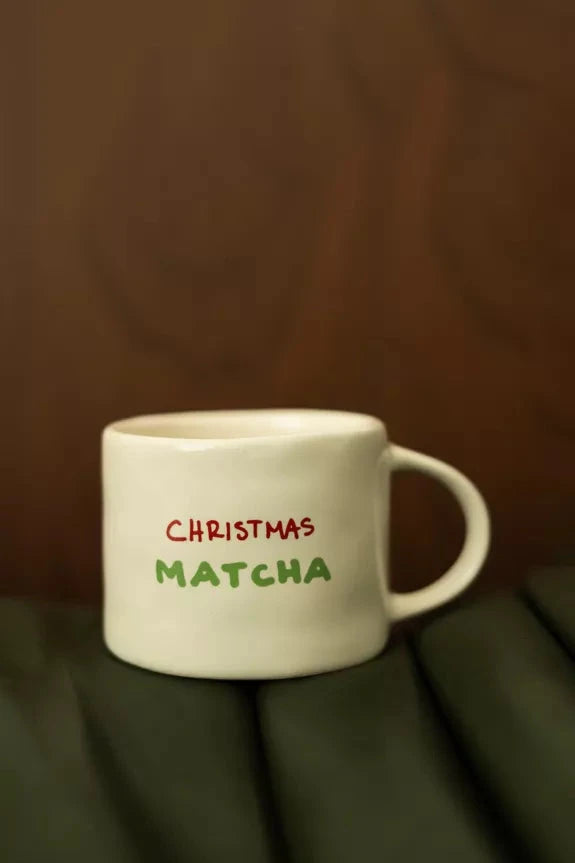 Mug Matcha de Noël en céramique blanche avec inscription 'Christmas Matcha' en rouge et vert, posé sur une surface en cuir vert foncé. Idéal pour boissons festives.
