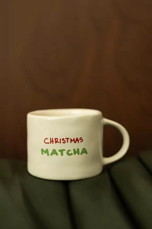 Mug Matcha de Noël en céramique blanche avec inscription 'Christmas Matcha' en rouge et vert, posé sur une surface en cuir vert foncé. Idéal pour boissons festives.
